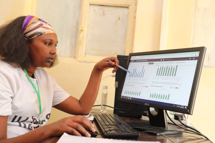 Lasha, seated at a desk, is gesturing with her pen as she refers to bar graphs on her computer monitor.