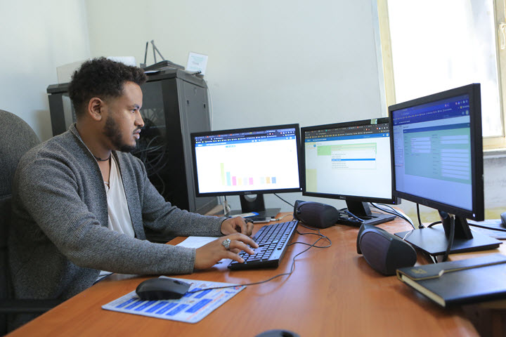Biniam is seated at a desk entering digital health information across three monitors