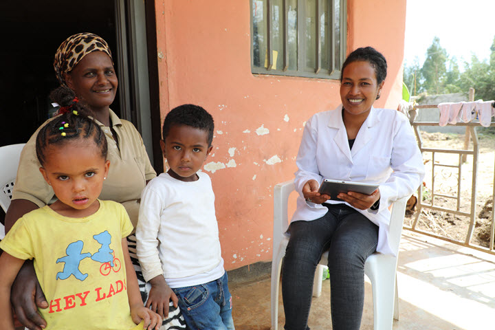 Freiwot is holding her tablet and smiling as she meets with a mother and two young children in front of their home.