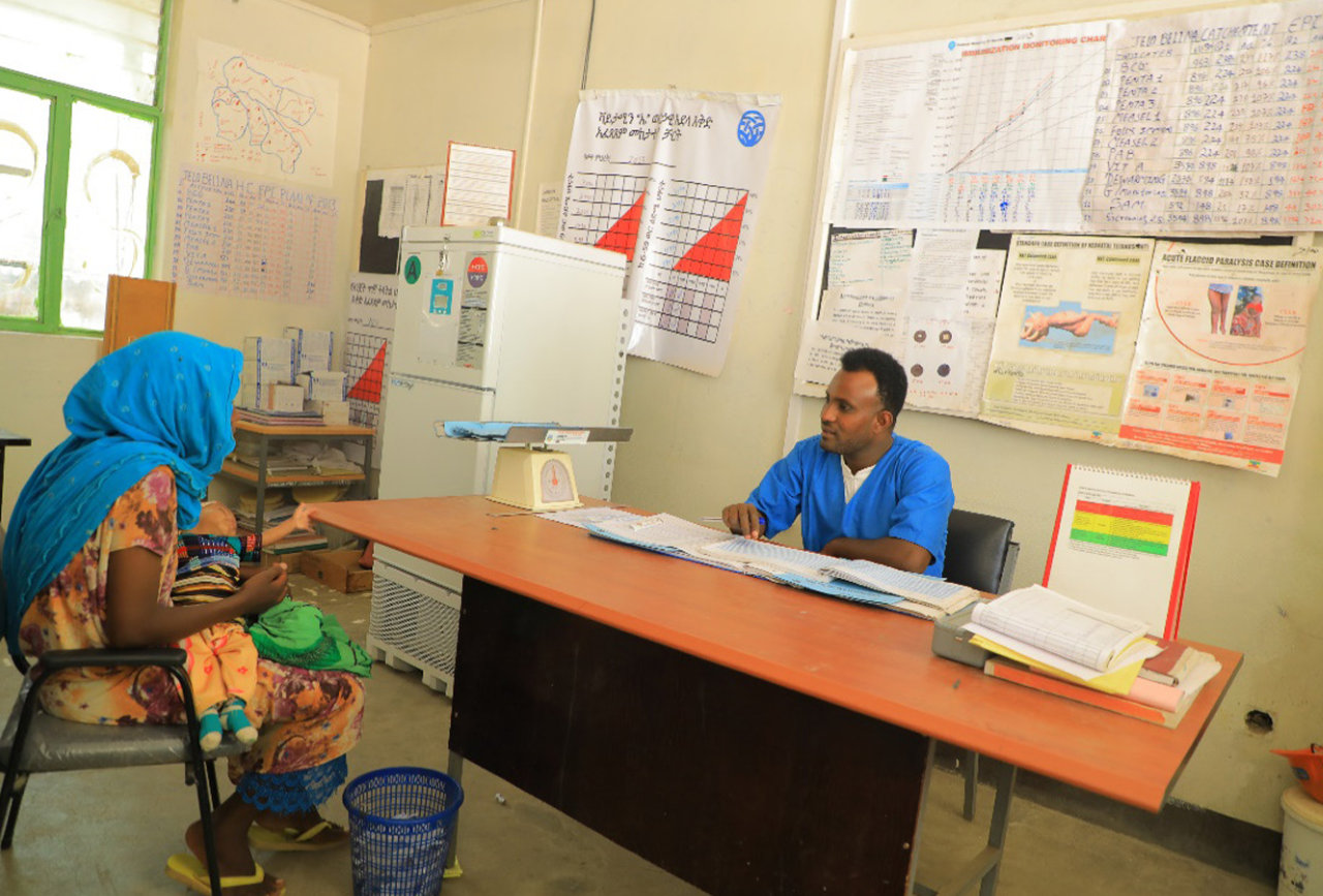Mother, with baby in her lap, in conversation with clinician who is consulting data while seated at his desk in the clinic.